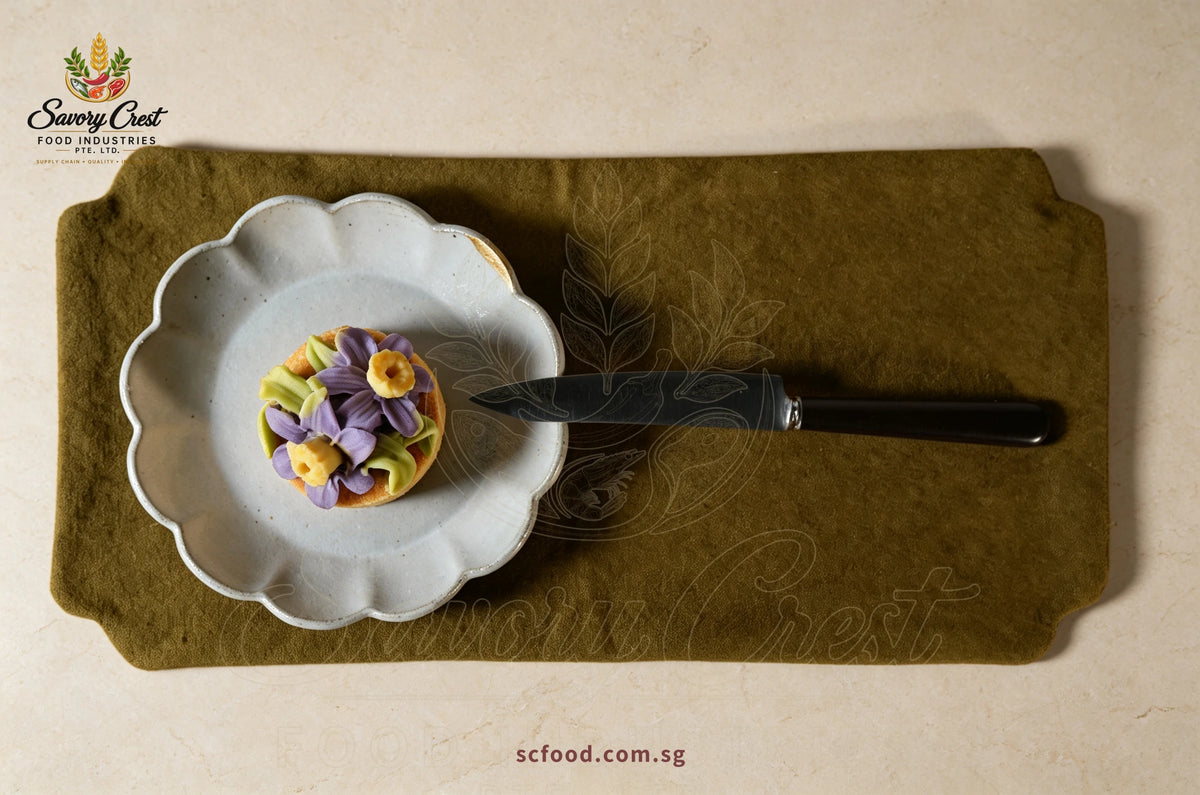 Overhead view of a purple floral pastry on a scalloped plate beside a knife on an olive-green table mat.